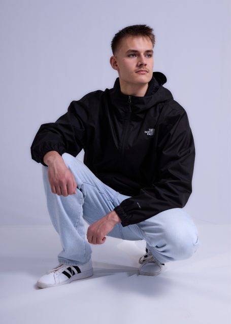 Junger Mann in schwarzer Jacke und hellblauen Jeans hockt auf einem neutralen Hintergrund.