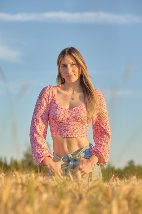 Junge Frau in einem Feld mit hohem Gras, lächelt und trägt ein buntes Oberteil.