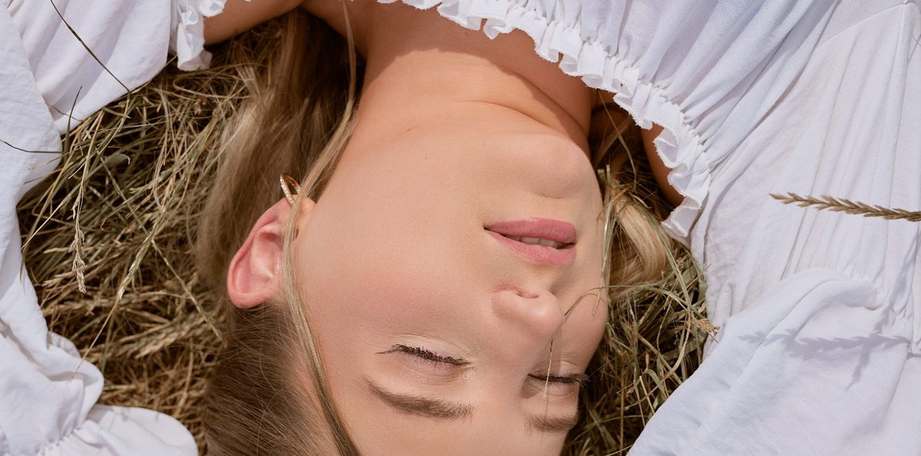 Frau mit geschlossenen Augen, liegend auf Heu, in einem weißen Oberteil.