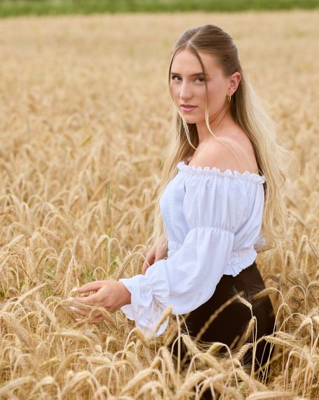 Junge Frau in weißer Bluse steht in einem goldenen Weizenfeld.