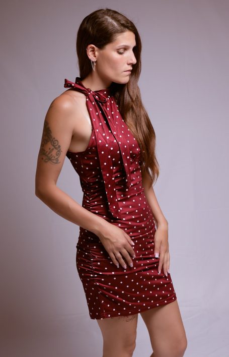 Frau in einem roten, gepunkteten Kleid mit Bindeband, seitlich stehend und nachdenklich.